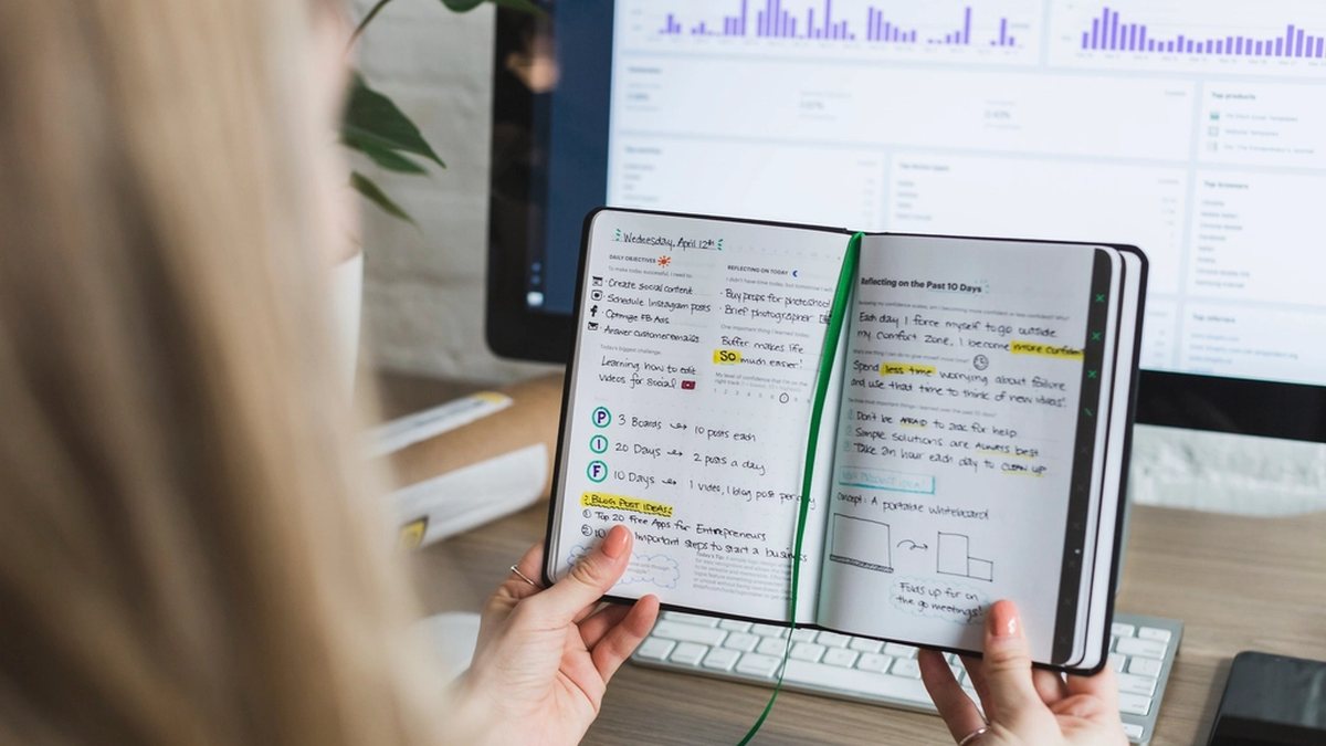 Persona hojeando una libreta de notas junto a una pantalla con gráficos