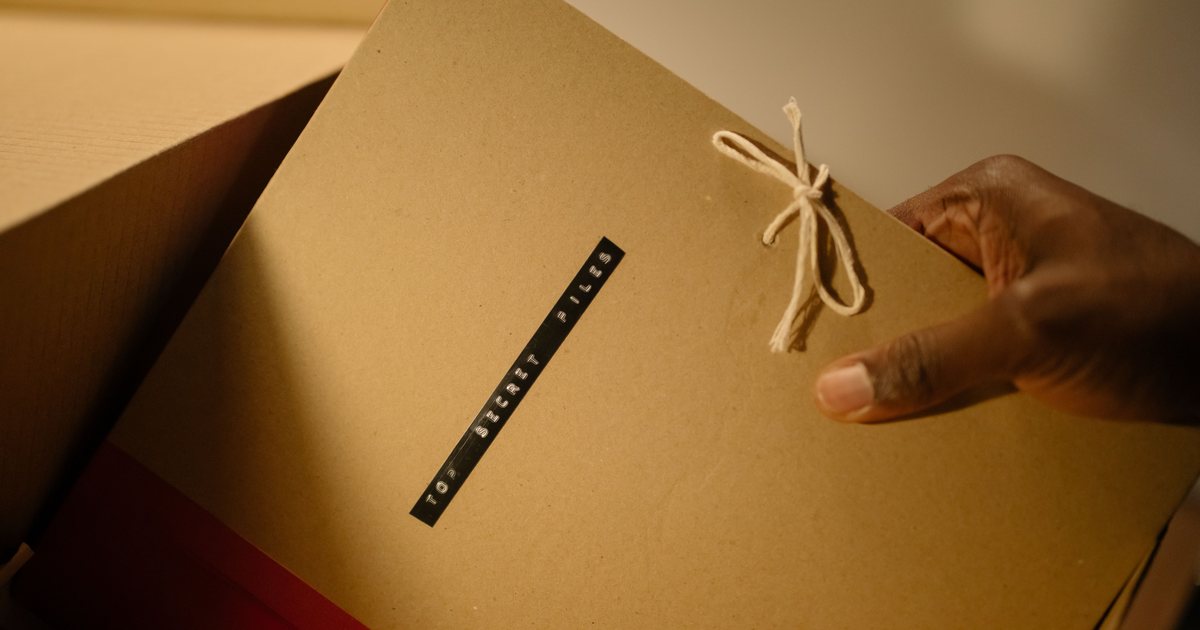 Una mano sostiene una carpeta de cartón cerrada con un cordón y una etiqueta que dice 'TOP SECRET FILES', asomando de una caja de archivo.