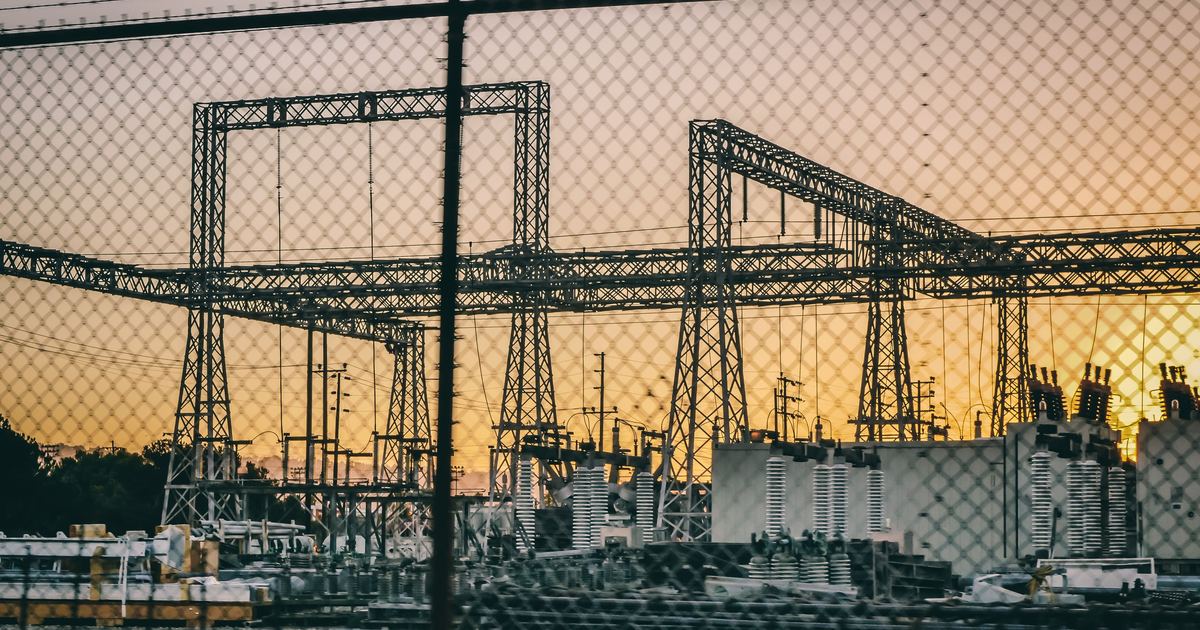 Subestación eléctrica industrial tras una valla metálica, con torres de alta tensión recortadas contra un cielo de atardecer.