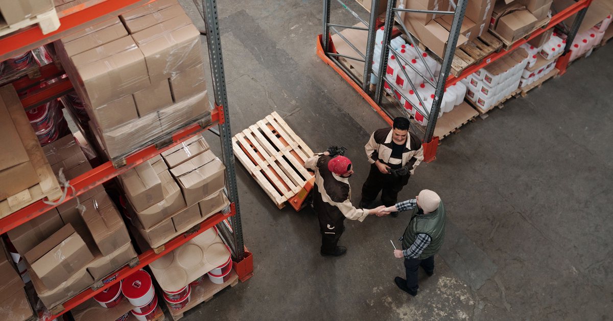 Vista aérea de un almacén logístico con estanterías llenas de cajas y bidones, donde tres operarios se dan la mano junto a una carretilla transportadora de palés