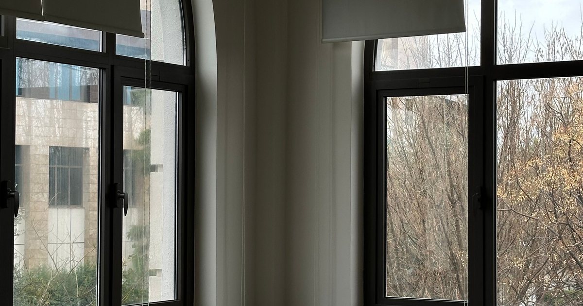 Un aula vacía con pupitres y sillas en fila, ventanas altas con luz natural y vistas a los árboles, sin ningún alumno presente.