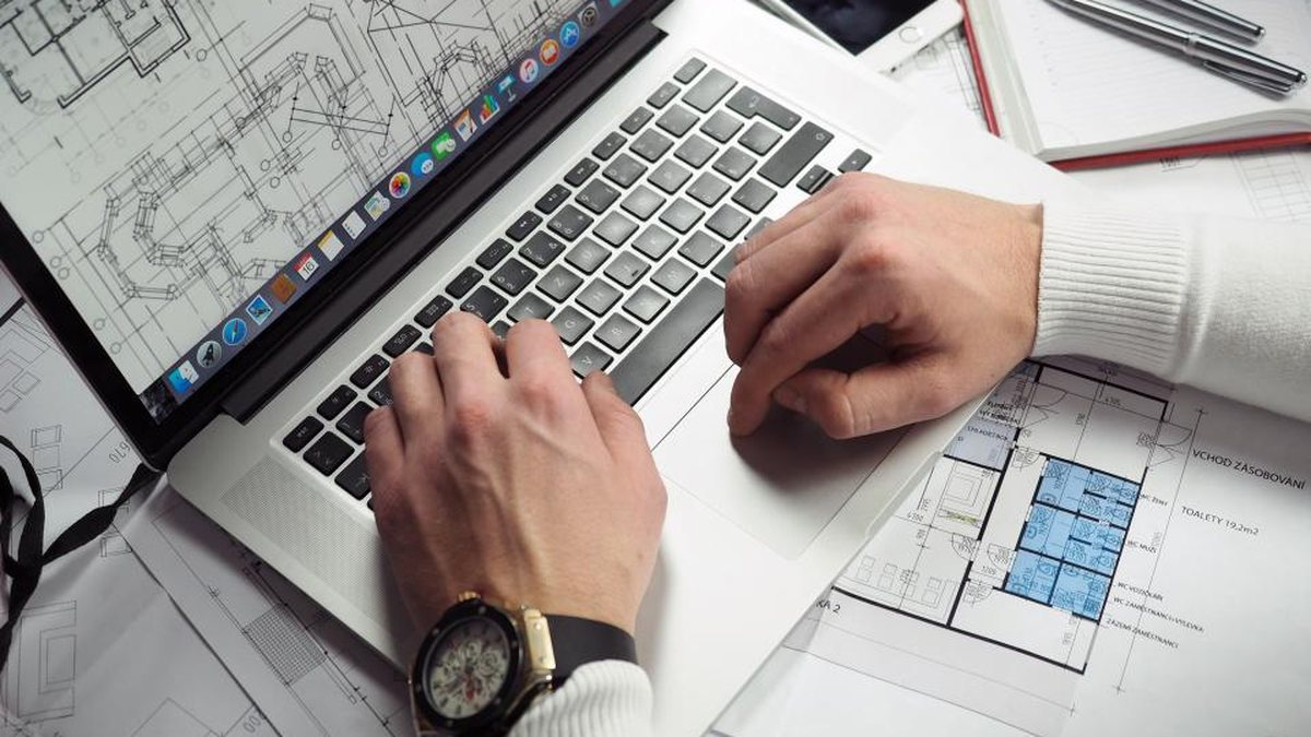 Manos escribiendo en un portátil junto a planos técnicos
