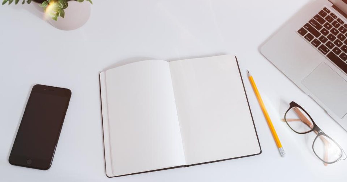 Portátil abierto junto a un cuaderno, un teléfono y unas gafas sobre una mesa blanca